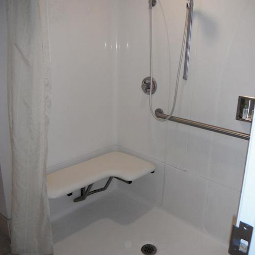 An accessible shower with a bench, handheld showerhead, grab bar, and drain on the floor. Shower curtain partially visible on the left side.