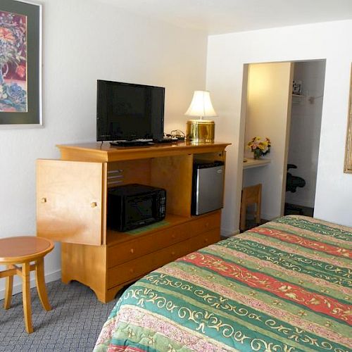 The image depicts a hotel room with a bed, wooden dresser with a TV, lamp, microwave, mini-fridge, and a small stool. A doorway is in the background.