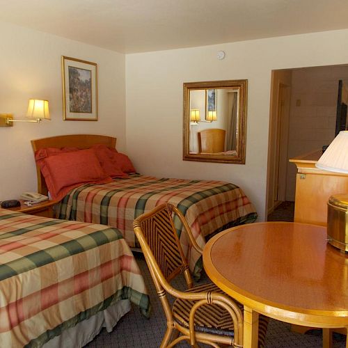 A hotel room with two beds, plaid bedding, a table with chairs, lamps, a TV, and a framed picture on the wall.