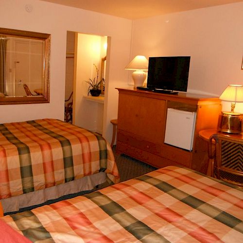 A cozy hotel room with two plaid-patterned beds, a TV, a mini-fridge on a wooden cabinet, and a warmly lit desk area with a chair and lamp.