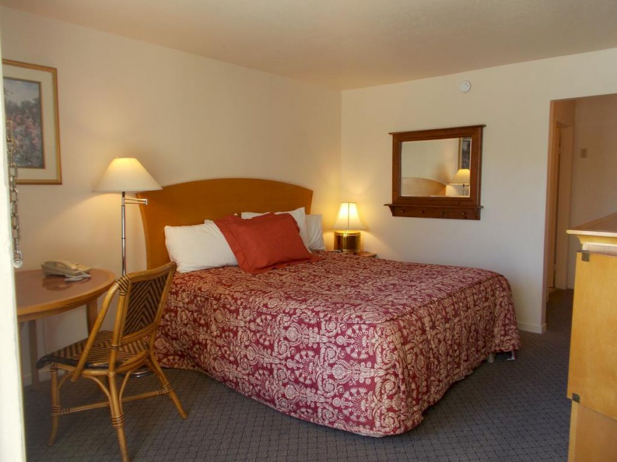 A neatly arranged hotel room featuring a king-size bed with patterned bedding, two bedside lamps, a wooden chair, a table, and a wall mirror.