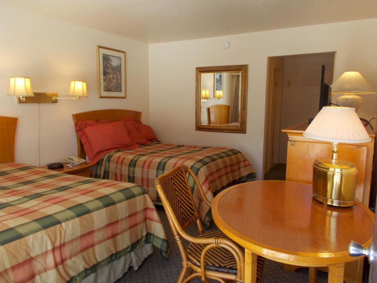 The image shows a hotel room with two beds, a table with two chairs, lamps, a dresser with a TV, and framed pictures on the wall.