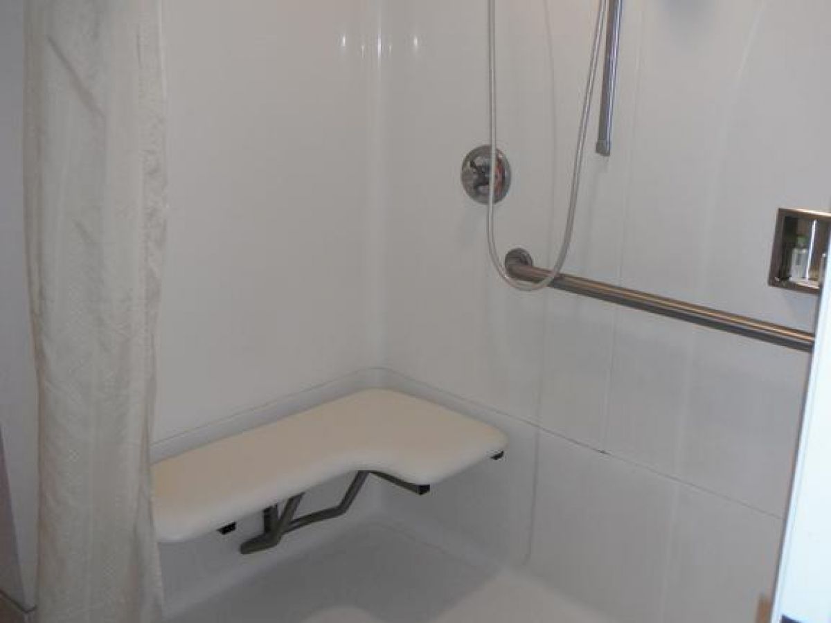 A shower area with a foldable bench, a handheld showerhead, a grab bar, and a shower curtain. The floor is tiled, and the walls are white.