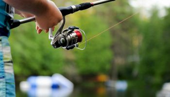 A person holding a fishing rod by a calm lake, with trees and blurred objects in the background.