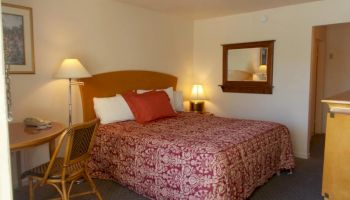 A cozy hotel room with a double bed dressed in red-patterned bedding, a small table with two chairs, a wall mirror, and warm lighting.