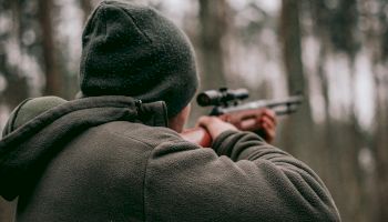 A person in outdoor clothing aims a rifle in a forest setting, with trees and foliage visible in the background.