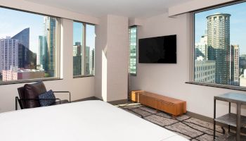 A modern hotel room with a large bed, flat-screen TV, armchair, and windows offering a cityscape view featuring tall buildings.
