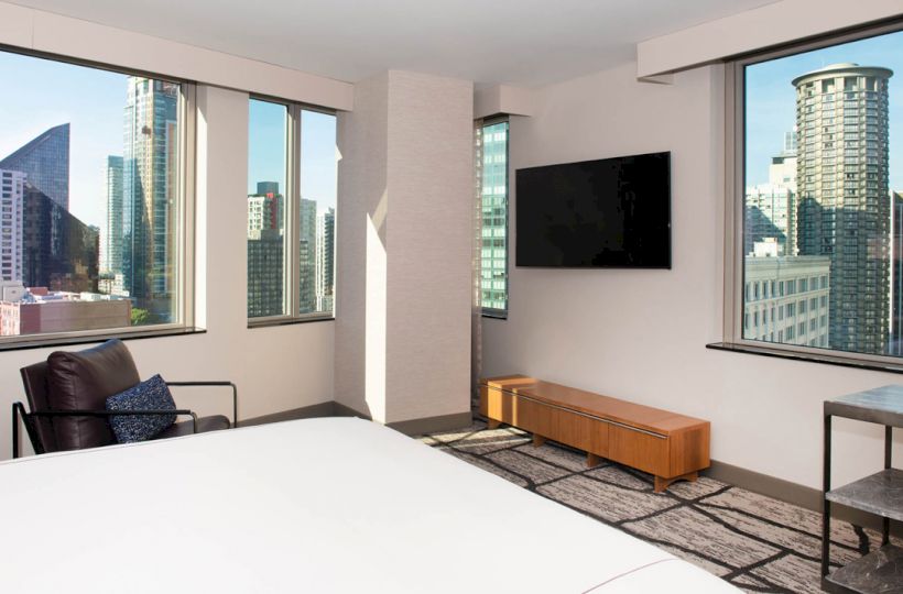 A modern hotel room with a large bed, flat-screen TV, armchair, and windows offering a cityscape view featuring tall buildings.