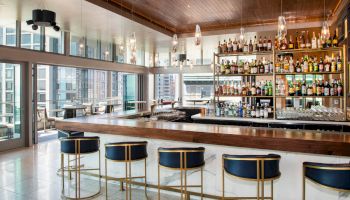 A stylish bar with a wooden counter, blue and gold bar stools, and many liquor bottles on the shelves, located in a modern high-rise building.