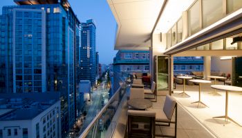 A modern cityscape at dusk viewed from a balcony, with chic outdoor seating and tables, adjacent to a well-lit indoor dining area.