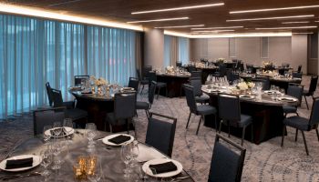 A banquet room is set up with round tables, black chairs, plates, glasses, napkins, and flower centerpieces, ready for an event or dinner.