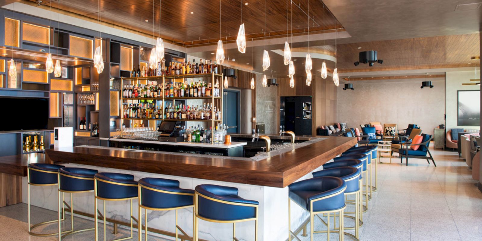 This image shows a modern bar with a long counter, blue bar stools, hanging light fixtures, and a well-stocked alcohol display, creating a cozy atmosphere.