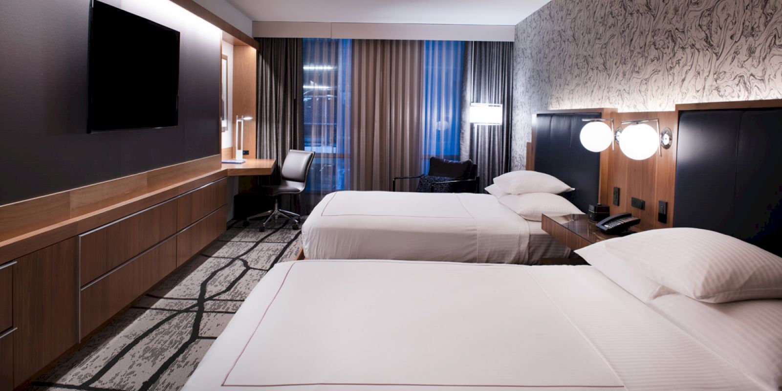 A modern hotel room features two neatly made beds, a flat-screen TV, a desk with a chair, a view outside through large windows, and contemporary decor.