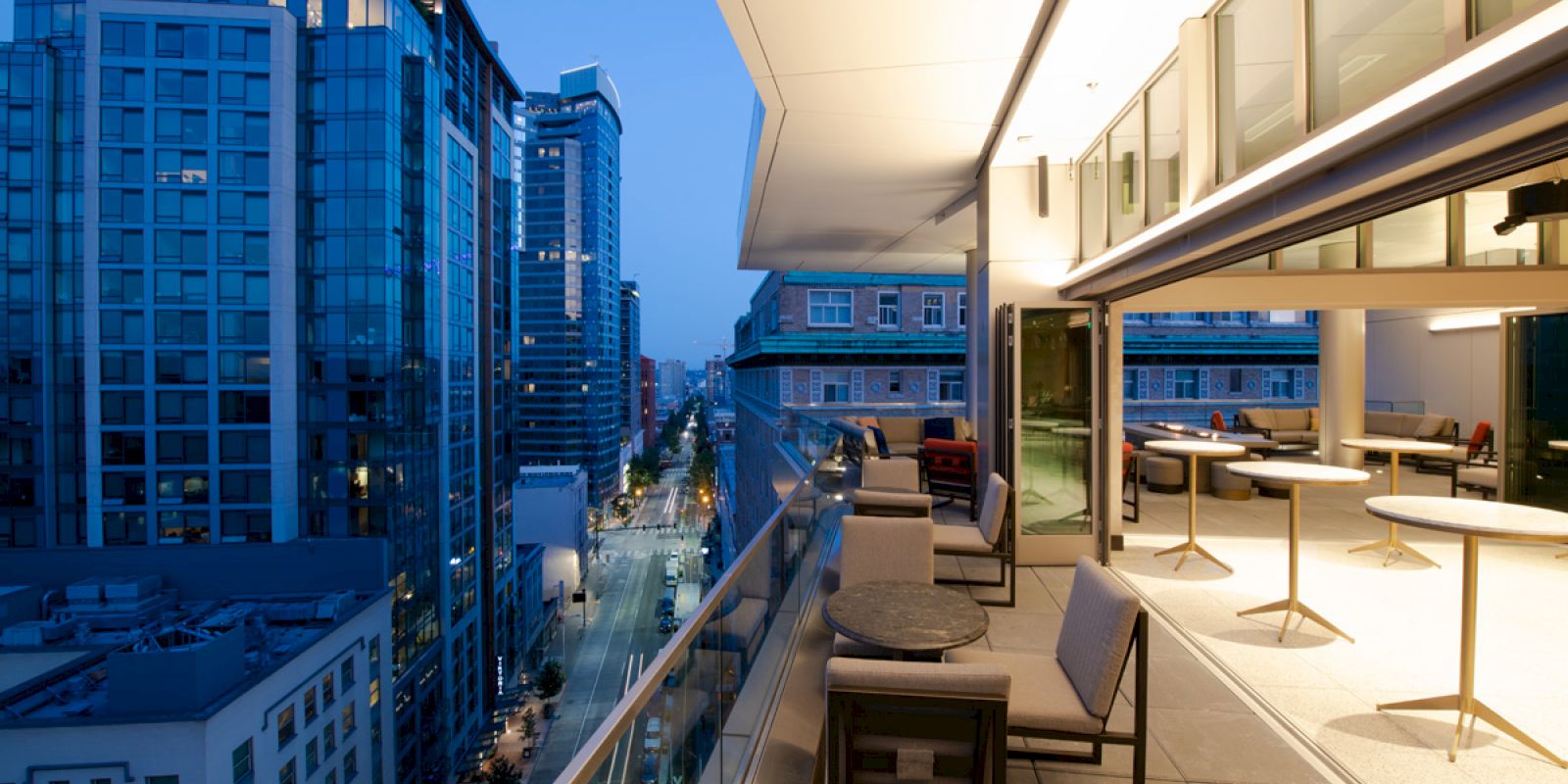 A modern cityscape at dusk with high-rise buildings and a spacious balcony featuring seating and tables, overlooking a street lined with buildings.