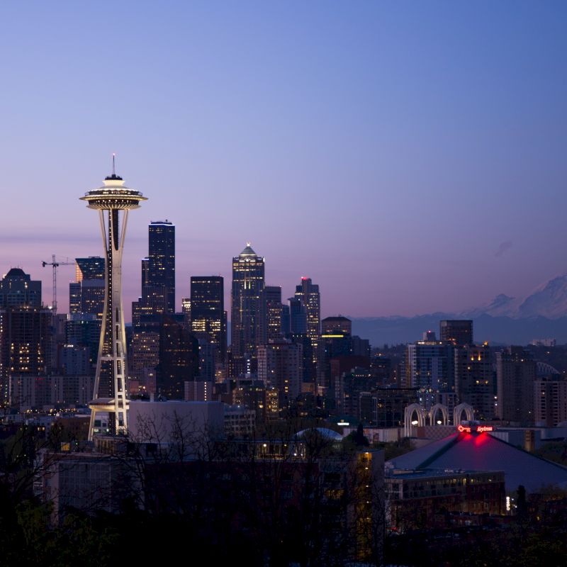 A cityscape of Seattle at dusk featuring the Space Needle, surrounding skyscrapers, and Mount Rainier visible in the distance against a twilight sky.