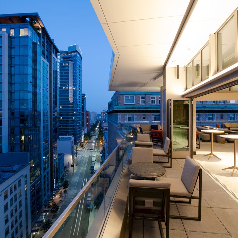 A modern cityscape viewed from a sleek, open-air balcony with seating and tables, overlooking tall buildings and a street below at dusk.
