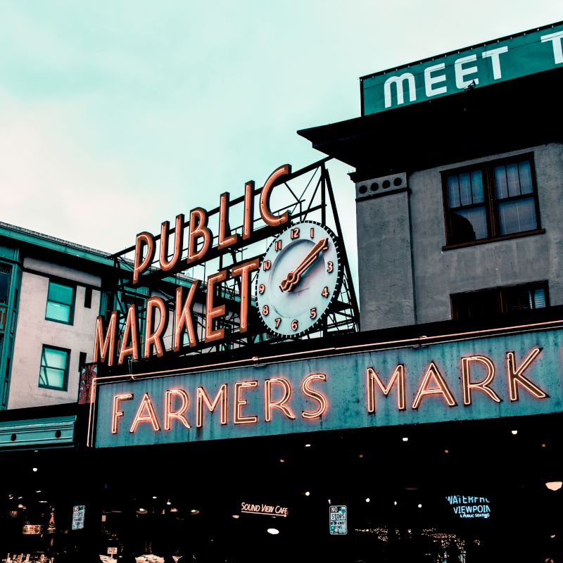 The image features neon signs for "Public Market" and "Farmers Market" on buildings, including a clock. It appears to be a busy urban market area.