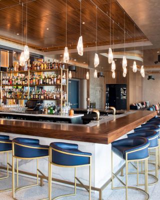 This image shows a stylish bar with modern lighting, a well-stocked bar counter, and blue barstools. There is a lounge area in the background.