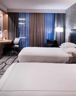 This image shows a modern hotel room with two double beds, a flat-screen TV, a desk with a chair, and wall art. The room has a clean, sleek design.