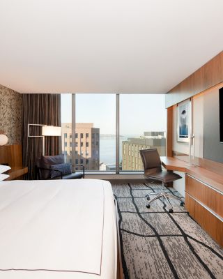 Modern hotel room with a large bed, wall-mounted TV, desk, chair, and floor-to-ceiling window offering a city view.