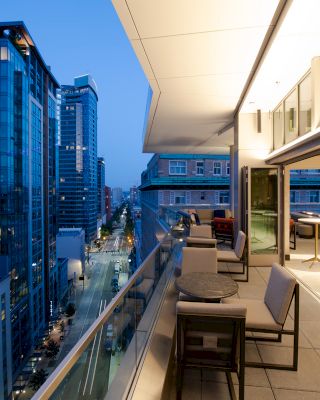 A modern cityscape during twilight, with high-rise buildings, an outdoor terrace featuring seating, tables, and streetlights below.