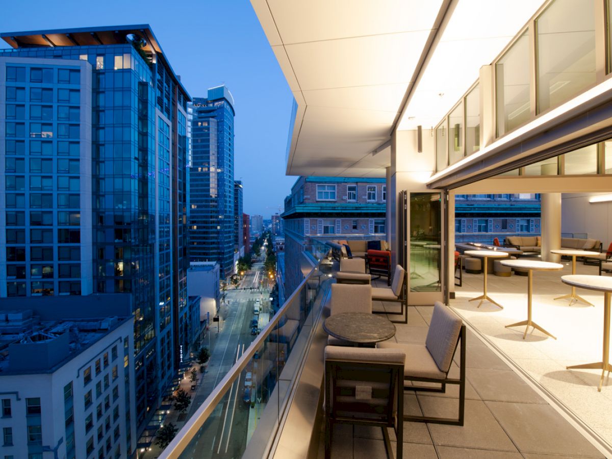 A modern rooftop terrace with seating and tables overlooks a cityscape of tall buildings at twilight, showcasing urban architecture.