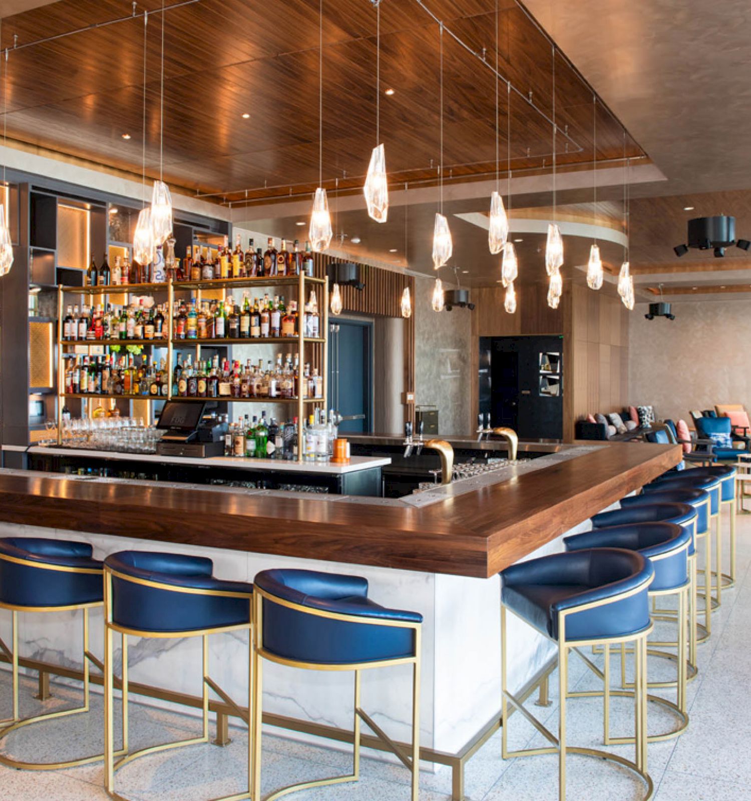 A modern bar with a well-stocked liquor display, blue stools, overhead pendant lights, and a spacious seating area in a stylish, well-lit environment.