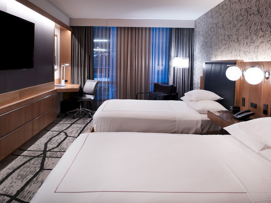 The image shows a modern hotel room with two beds, a flat-screen TV, a desk with a chair, and a large window with curtains.