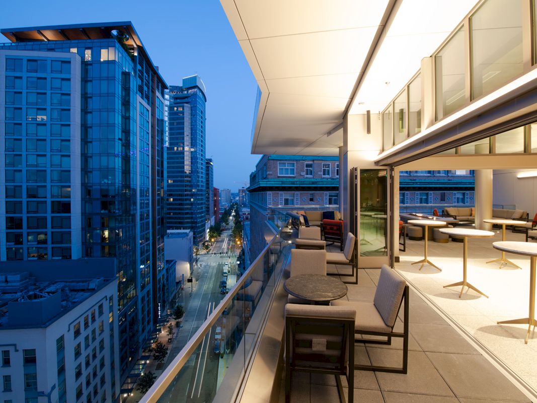 A modern cityscape at dusk with tall buildings and a well-lit balcony featuring tables and chairs, overlooking a street lined with lights.