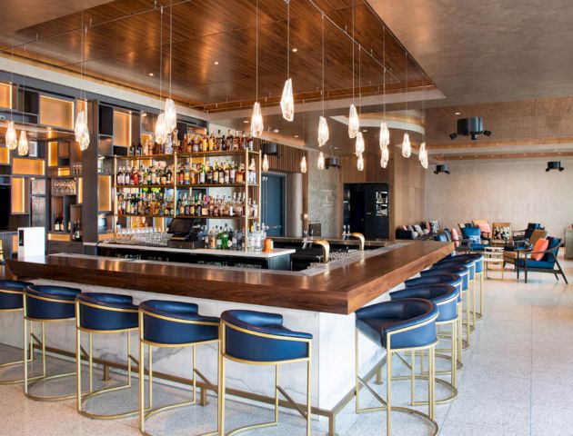 A modern bar with a wooden countertop, blue barstools, a variety of alcohol bottles, hanging pendant lights, and seating areas in the background.
