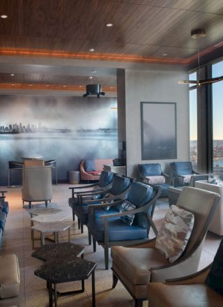 The image shows a modern lounge with comfortable seating, a wall featuring art of a skyline, and large windows with a view of a waterfront and a Ferris wheel.