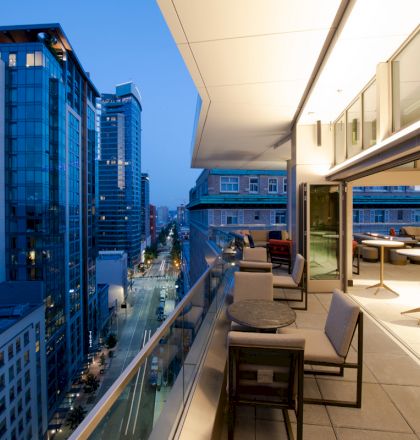 A modern cityscape at dusk with tall buildings and a lit-up balcony featuring tables and chairs overlooking the street below.