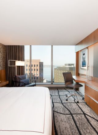 A modern hotel room with a large bed, wall-mounted TV, desk, chair, and a view of tall buildings through a large window, and natural light.