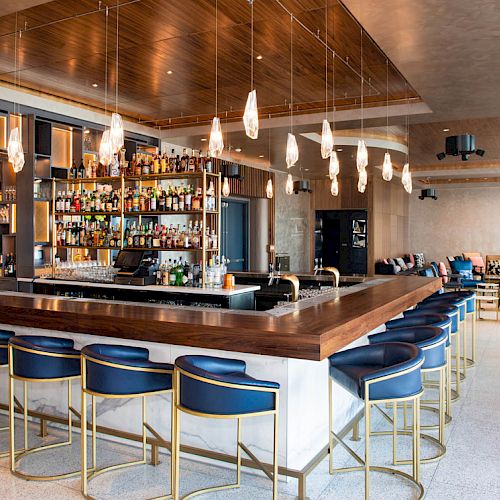 A modern bar with a wooden counter, blue bar stools, hanging lights, and a well-stocked shelf of liquor bottles. It has a bright, stylish interior.