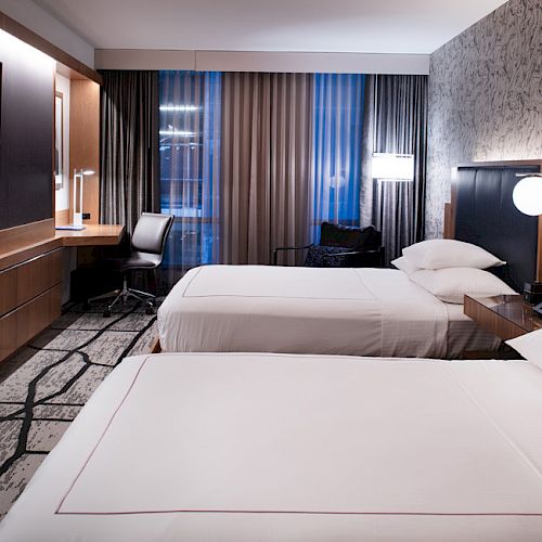 The image shows a modern hotel room with two neatly made beds, a wall-mounted TV, a desk chair, and stylish lighting fixtures.