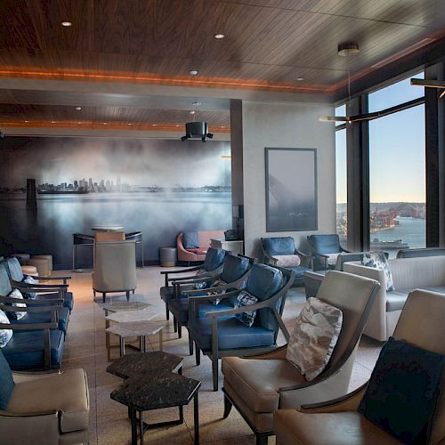 A modern lounge area with chairs and tables, large windows offering a view of a ferris wheel and water, and a wall mural depicting a cityscape.