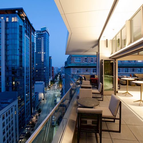A modern cityscape at dusk, seen from a rooftop terrace with tables and chairs, alongside tall, illuminated buildings.