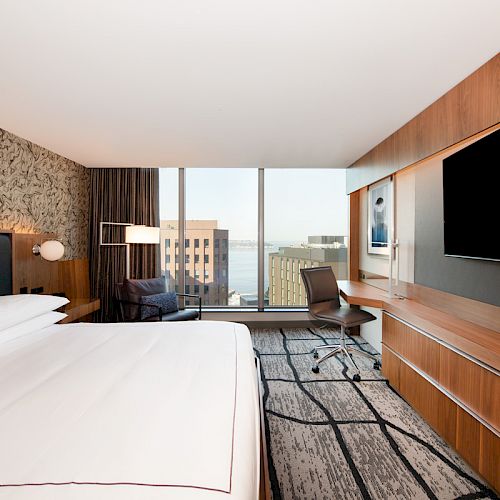 A modern hotel room with a large bed, TV, desk, chair, and a sofa with a window view of buildings and water in the distance.