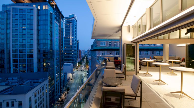 The image shows a modern high-rise cityscape at dusk, with a balcony featuring seating and tables overlooking a busy urban street.
