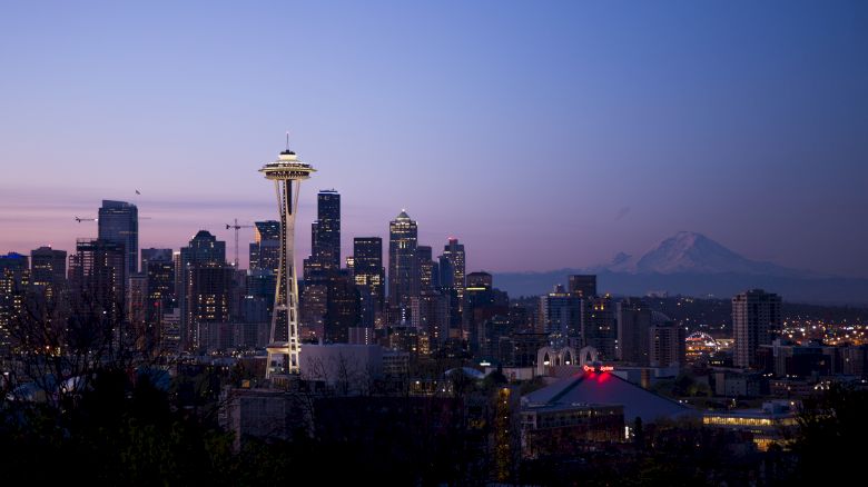 A cityscape at dusk featuring the iconic Space Needle, surrounded by skyscrapers, with a distant mountain visible against the twilight sky.