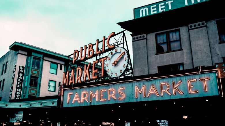 This image shows a public farmers market with neon signs, a clock, and surrounding buildings, likely indicating a popular urban shopping area.