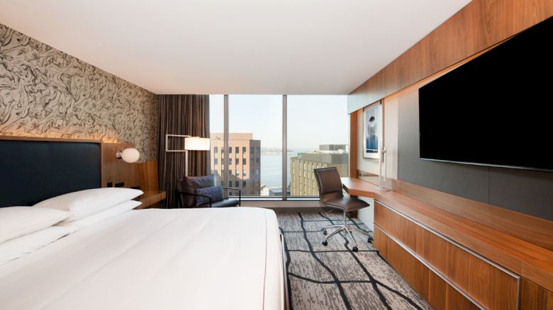 A modern hotel room with a large bed, a wall-mounted TV, a desk, and a chair, featuring a large window with a cityscape view.