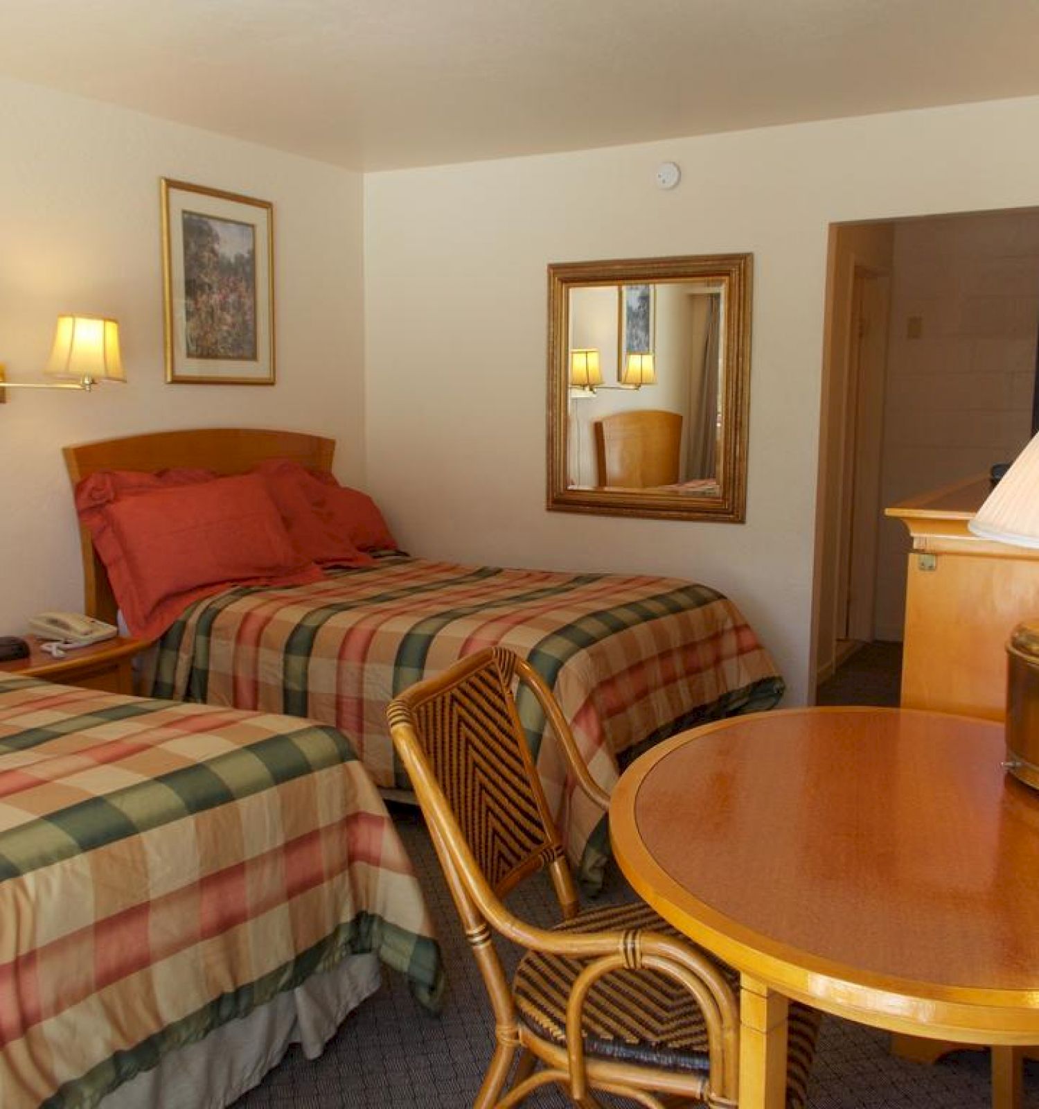 The image shows a hotel room with two double beds, plaid bedspreads, a wooden table, two chairs, a TV, a lamp, and framed artworks on the walls.