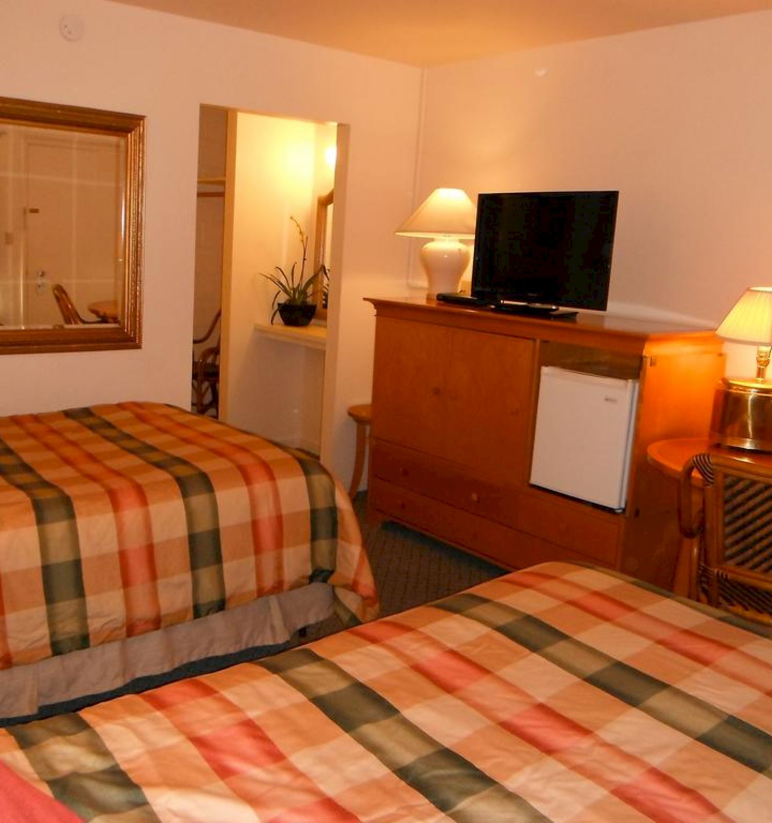 The image shows a cozy hotel room with two beds, a TV atop a dresser, a chair, a table lamp, a wall mirror, a picture, and welcoming lighting.