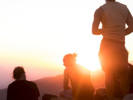 Three people are sitting on rocks, watching the sunset over mountains. The scene has a warm, serene atmosphere.