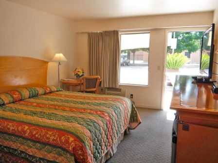 A hotel room with a bed, a small table with chairs, a TV on a dresser, and a window with curtains. The decor is simple and functional.